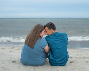 Couple sitting on beach facing ocean, leaning heads together romantically, wearing blue outfits on cloudy day.