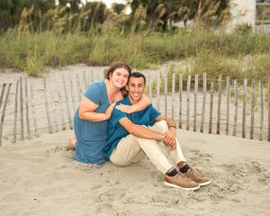 Couple sitting together on sandy beach by dune fence, wearing teal and khaki outfits, smiling warmly.