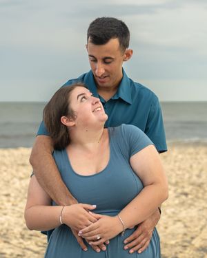 Couple embracing on a sandy beach, woman in blue dress looking up lovingly at man in teal shirt, ocean in background.