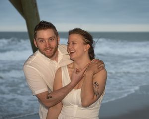 Happy couple embracing on a beach near a pier, woman in white dress laughing, man in white shirt, ocean waves behind them.