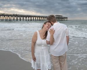 Couple in white clothing embracing on a beach at sunset with a pier in the background.
