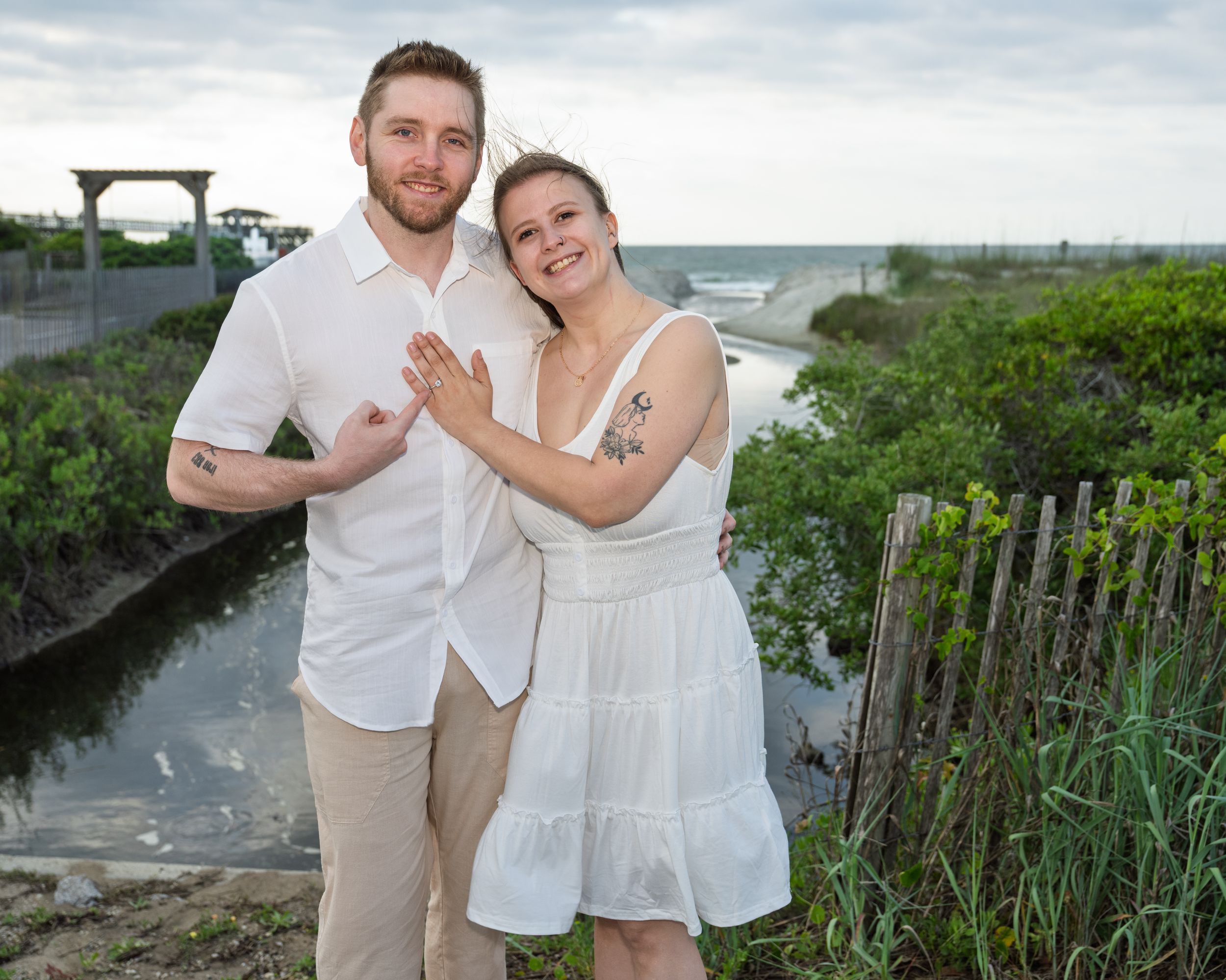 Happy couple in white outfits posing outdoors near a waterway with lush greenery and a pergola in the background.