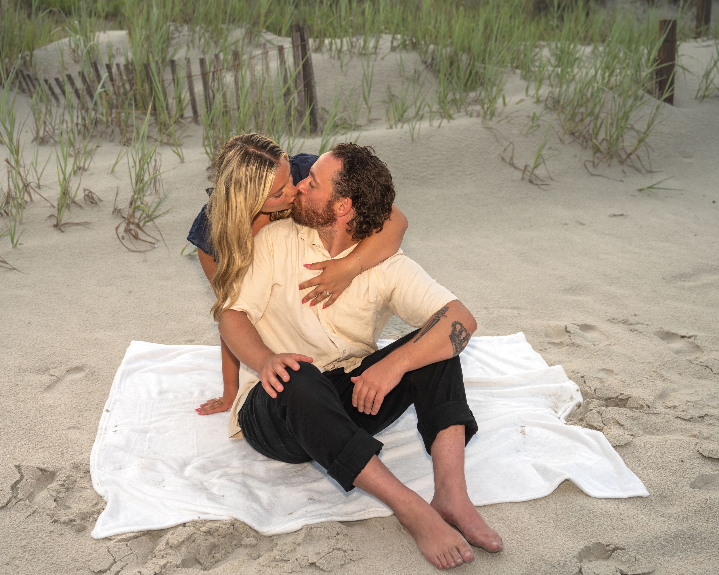 Couple kissing romantically on a white blanket on a sandy beach with dunes and grass in the background.