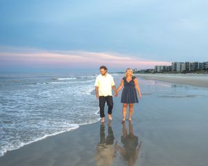 Couple holding hands walking barefoot on a sandy beach at sunset with ocean waves and coastal buildings.