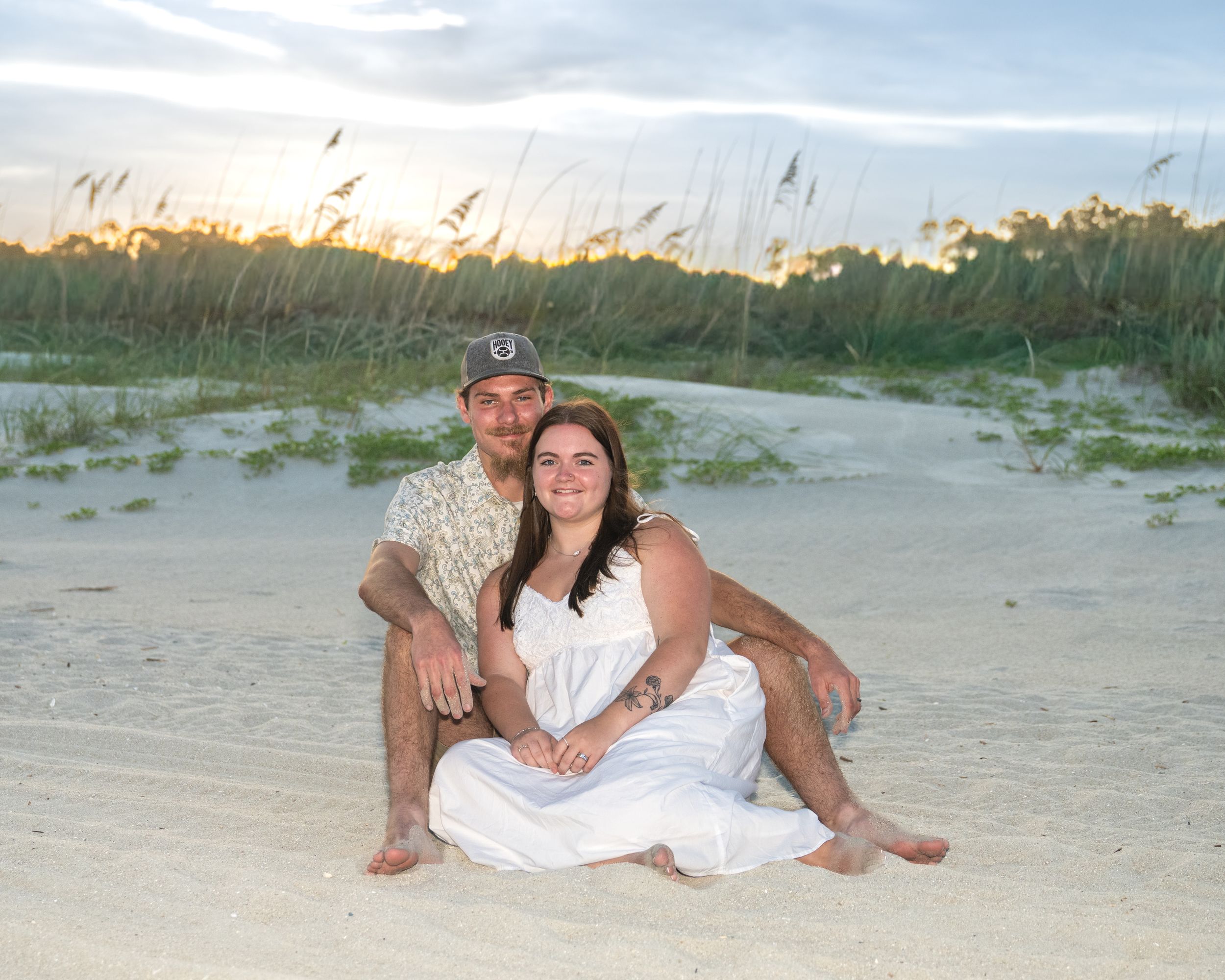 Couple sitting together on a sandy beach at sunset, woman in white dress, man in floral shirt, sea oats in background.