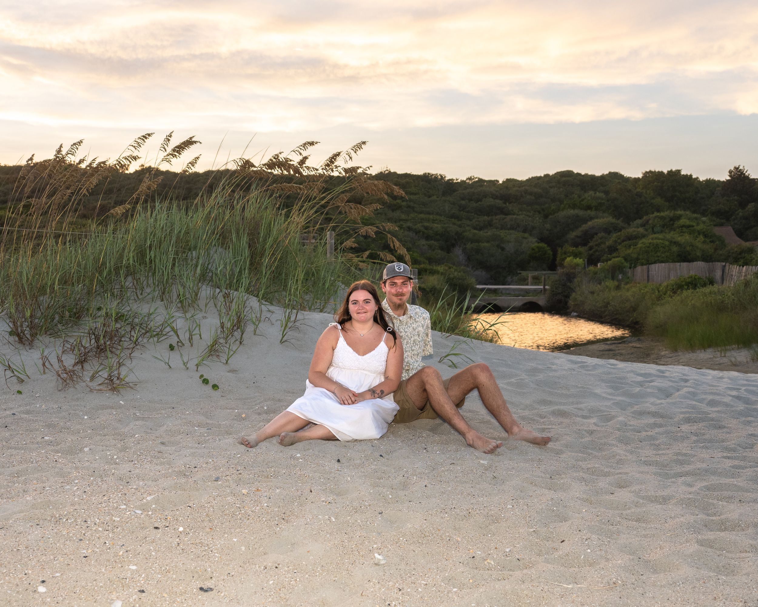 Couple sitting together on sandy beach dunes at sunset, woman in white dress, man in floral shirt, coastal grasses behind them.