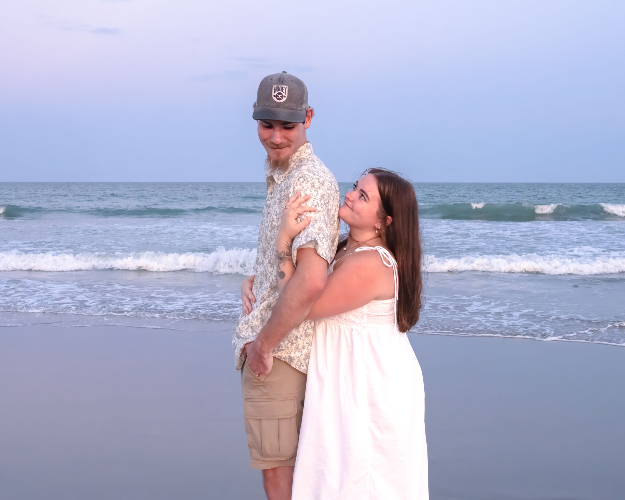 Couple posing romantically on a beach at dusk, man in floral shirt and cap, woman in white dress by ocean waves.