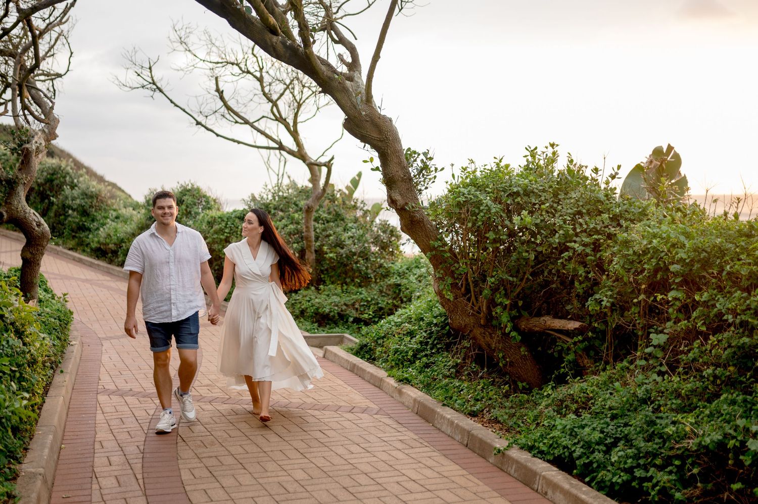 Couple walking hand-in-hand on a brick path surrounded by lush greenery at sunset.