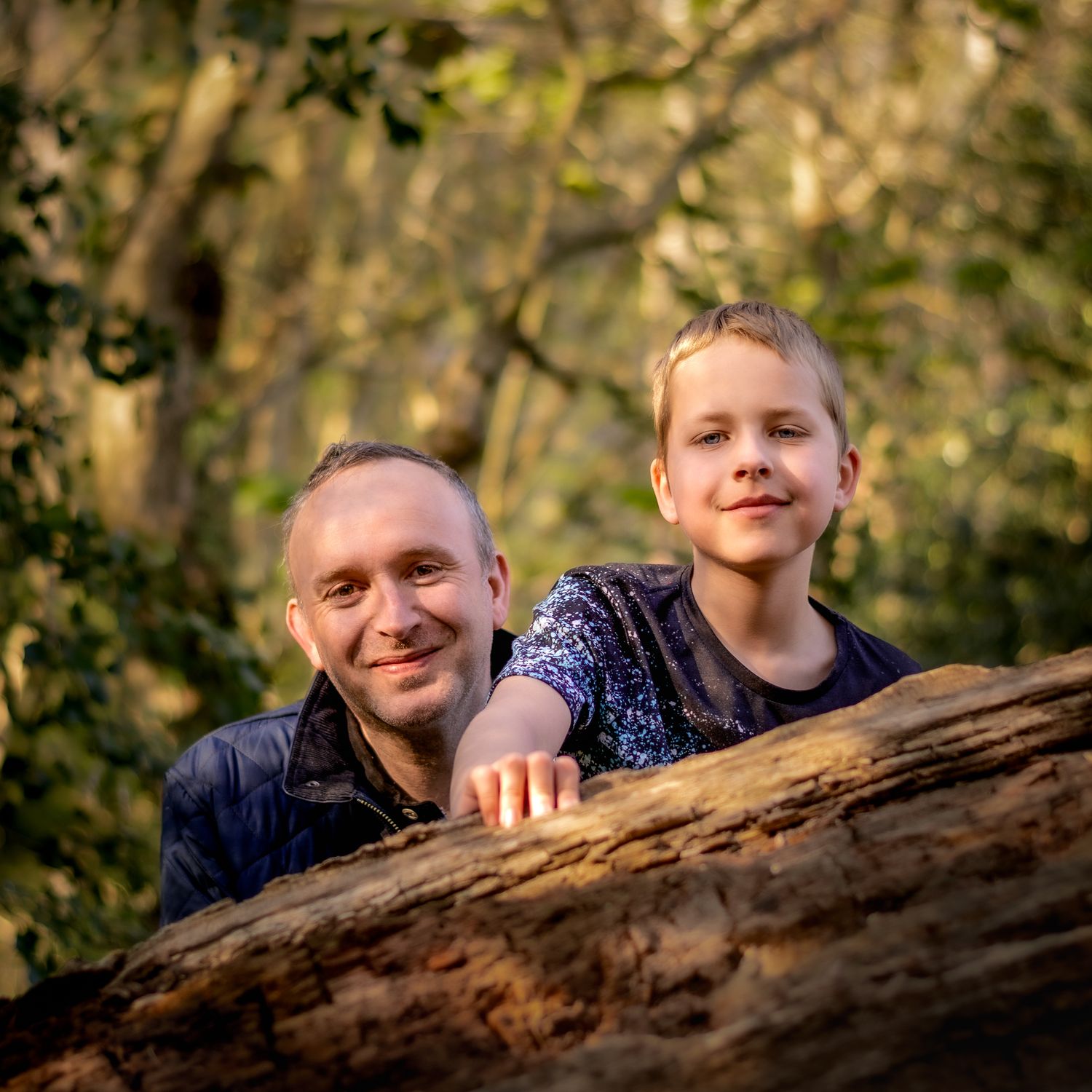 A man and young boy smiling behind a fallen log in a sunlit forest setting.