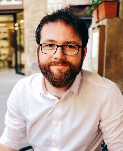 Professional in glasses and beard smiles warmly while sitting in a bright modern office space.