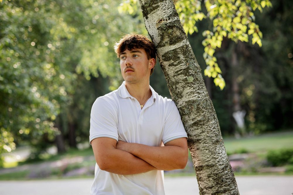 Young man with crossed arms leaning against a tree in a sunny park, wearing a white polo shirt.