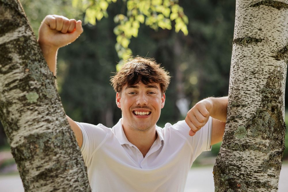 Happy young man smiling between two birch trees in a park, raising fists in celebration outdoors.
