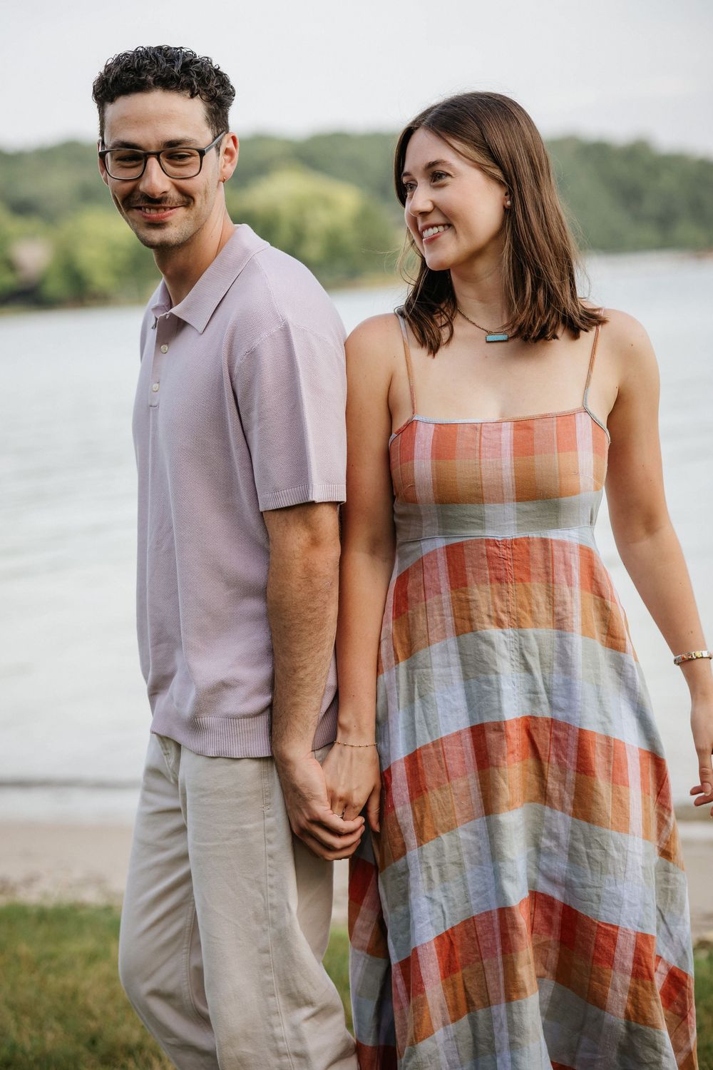 A young couple holds hands and smiles near a lake, wearing casual summer outfits.