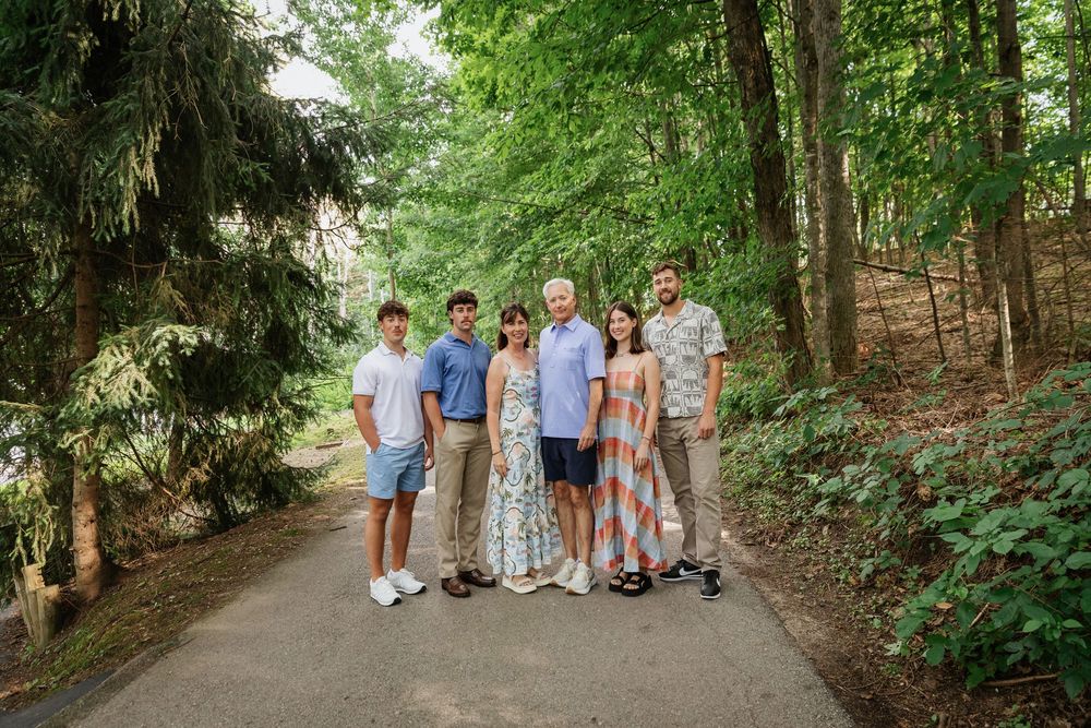 Family of six posing together on a tree-lined path surrounded by lush green foliage on a sunny day.