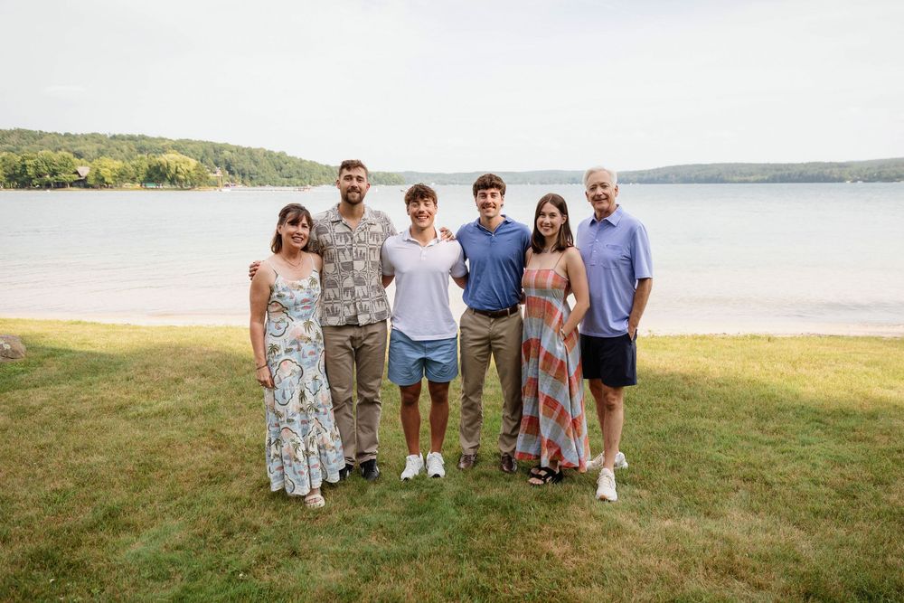 Group of six people smiling together on a grassy lakeside lawn with trees and water in the background.