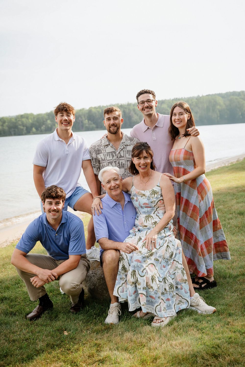 Family of seven posing outdoors by a lake, smiling together on a green lawn on a warm summer day.