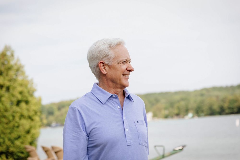 Smiling senior man with gray hair wearing a blue shirt, standing outdoors near a lake with trees in the background.