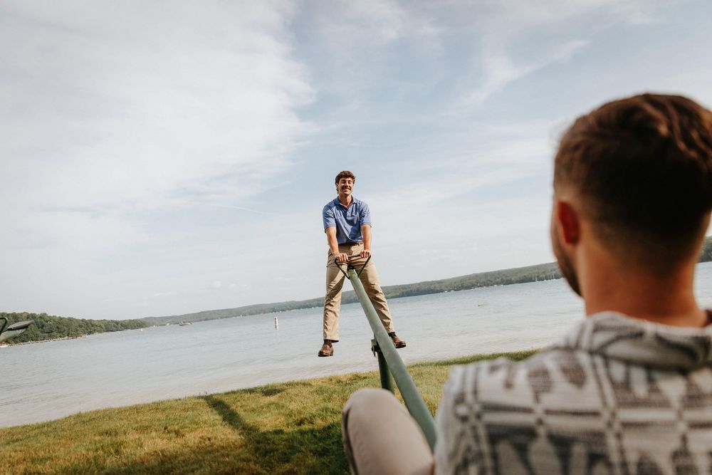 Two men playing a lawn game near a lakeside, one standing ready to catch on a sunny day.