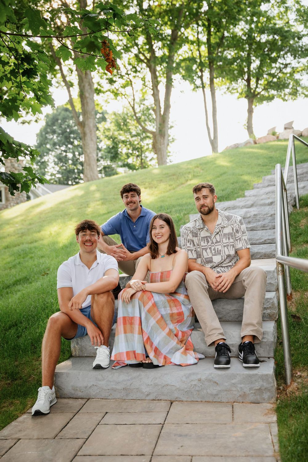 Four young friends smiling and posing together on outdoor stone steps surrounded by lush green trees and grass.