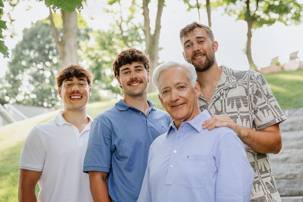 Four smiling men of different ages pose together outdoors in a lush green park setting on a sunny day.
