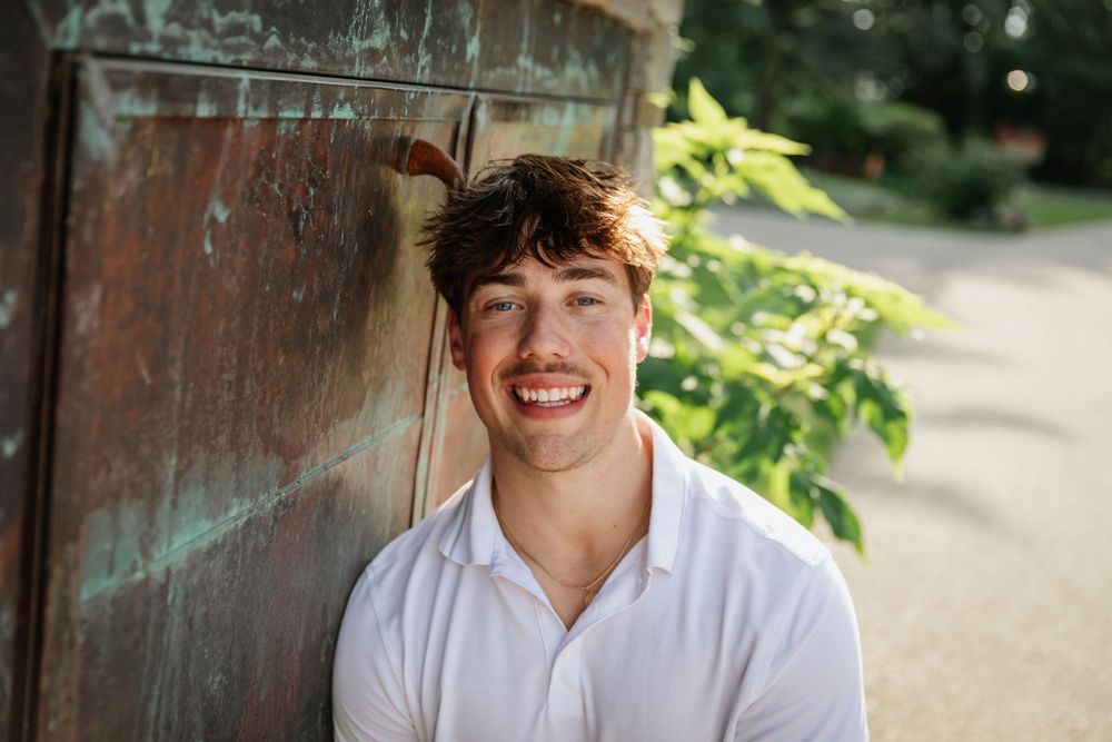 Smiling young man with brown hair leaning against a rustic wooden door outdoors in natural light.