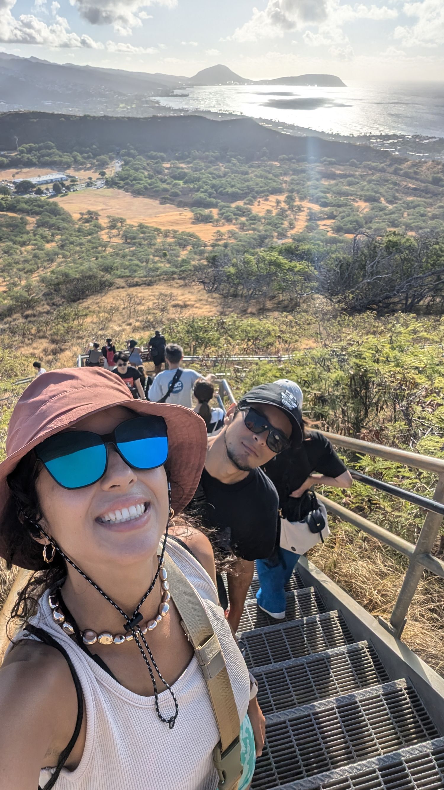Two hikers take a selfie on metal stairs at Diamond Head crater with panoramic Hawaii coastline views behind them.