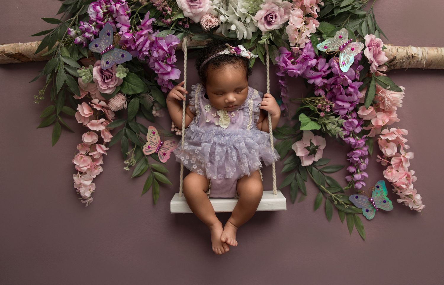 A baby rests on a rustic wooden bench surrounded by pink and purple floral arrangements.