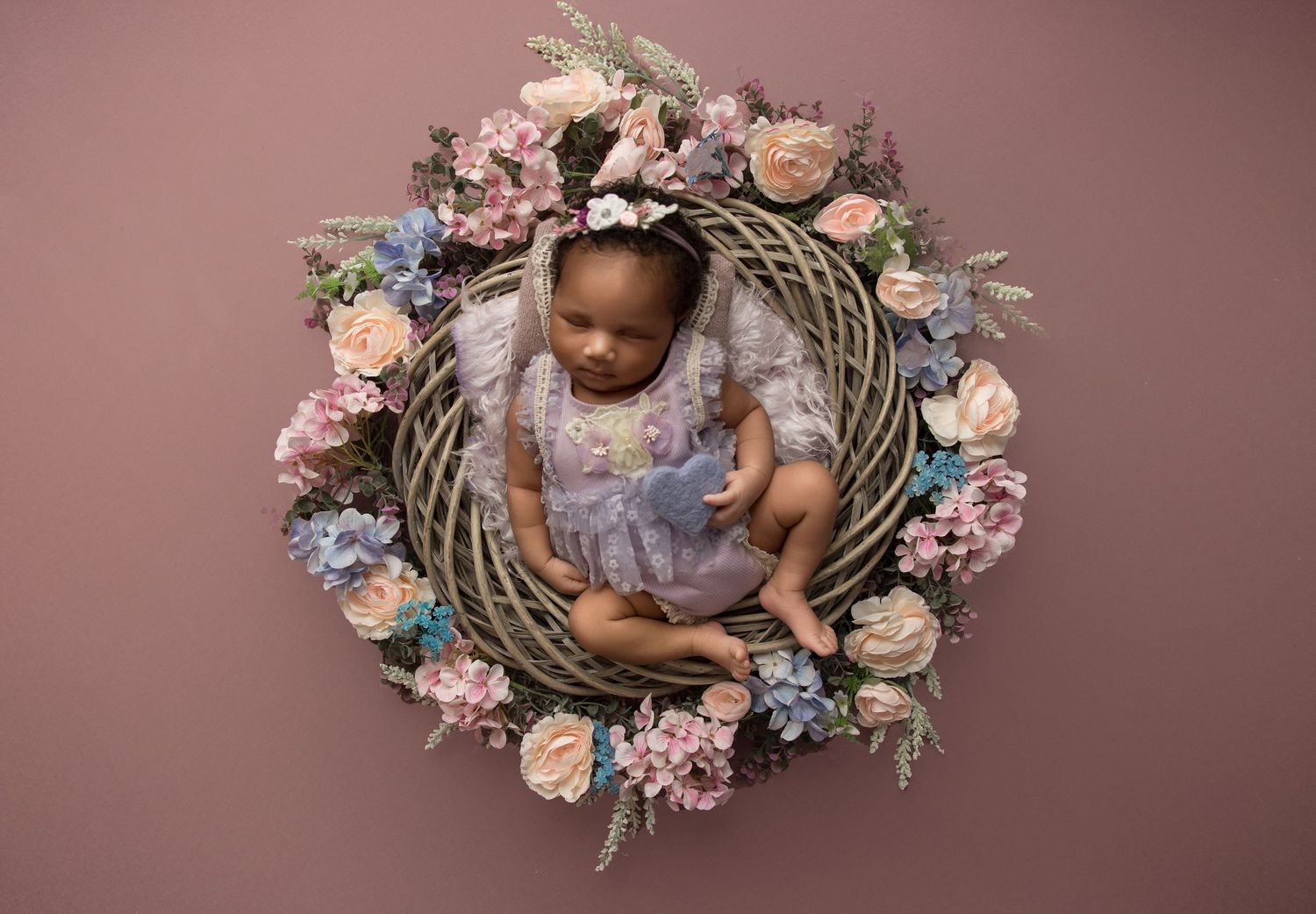 Newborn baby sleeping in a floral wreath nest with pink and peach roses on a mauve backdrop.
