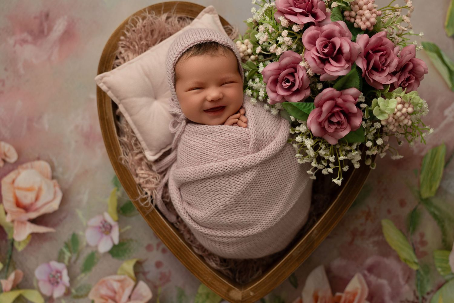 A newborn wrapped in soft gray swaddle rests in a heart-shaped basket with pink roses and floral backdrop.