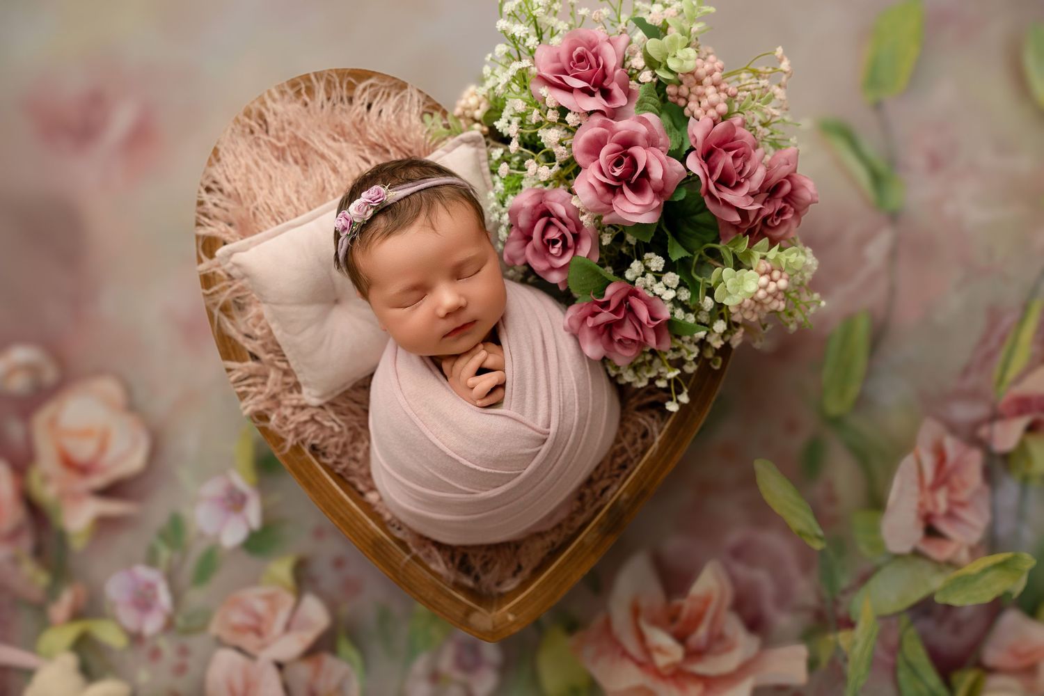 Sleeping newborn in heart-shaped basket with pink roses and baby's breath flowers against floral background.
