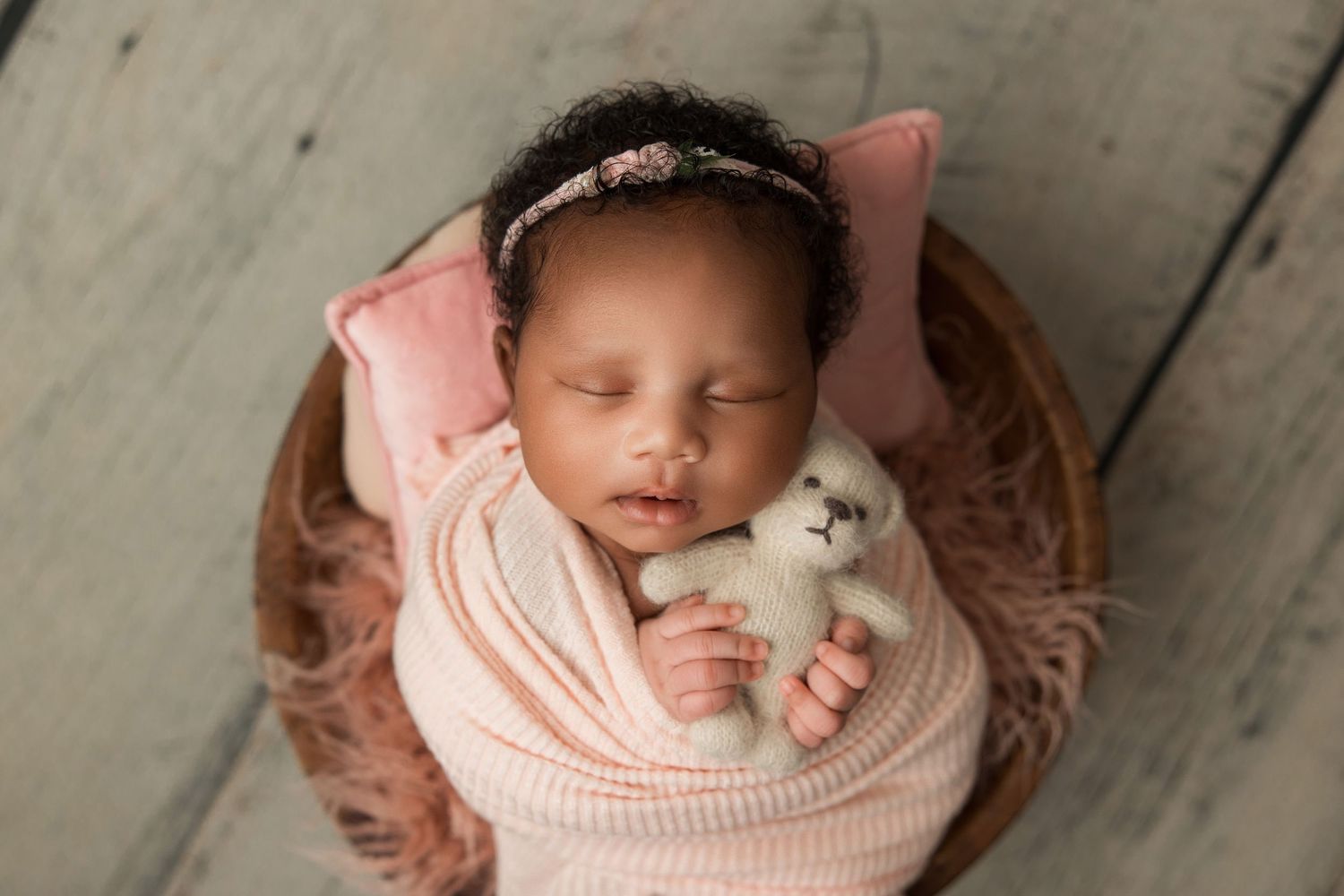 A sleeping newborn baby wrapped in soft pink fabric lies peacefully in a cozy wooden basket.