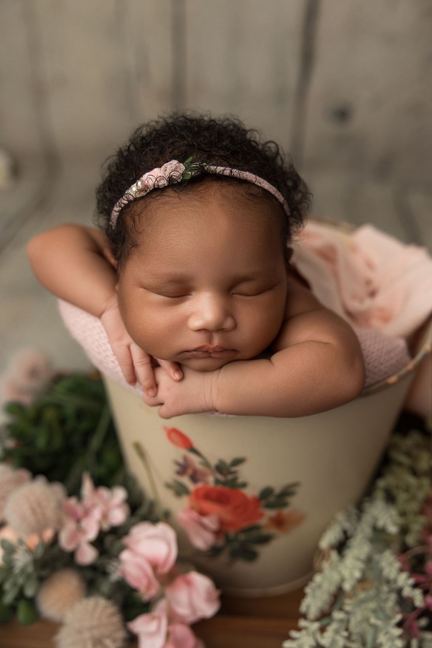 Sleeping newborn rests peacefully in a floral teacup surrounded by pink flowers and greenery in a dreamy studio setting.