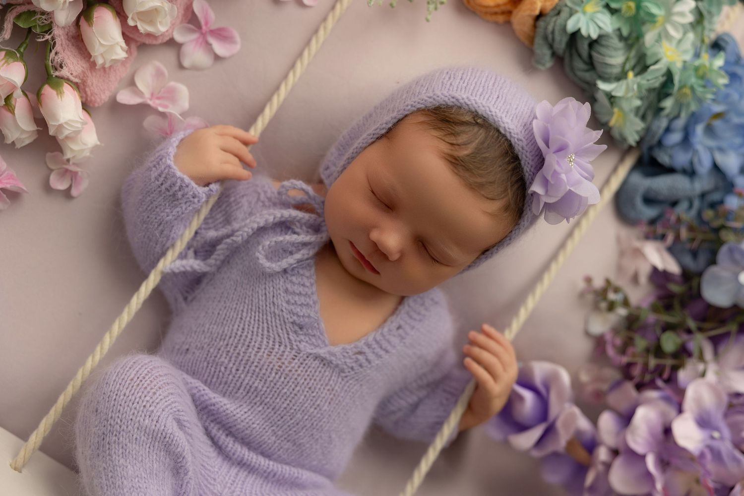 Newborn photography setup with purple knit blanket and delicate floral arrangements.