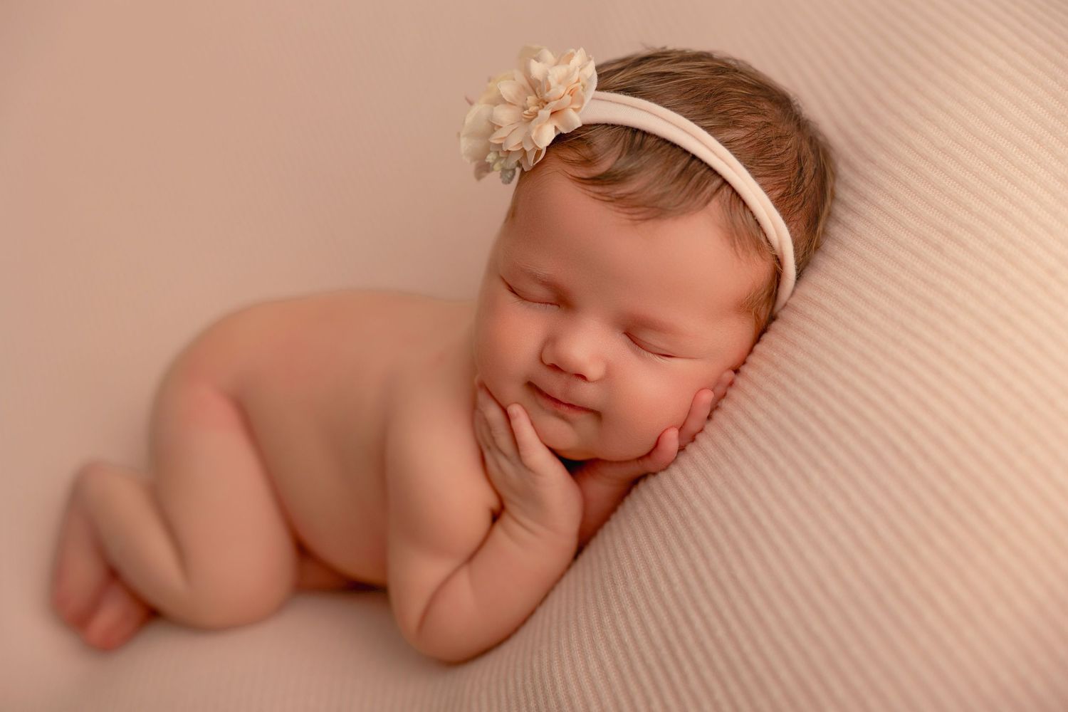 A sleeping newborn baby wearing a headband rests peacefully on a textured beige blanket.