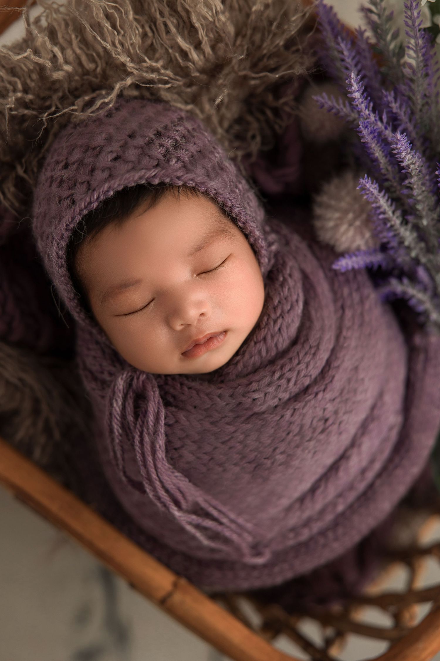 Sleeping baby wrapped in soft purple knitted blanket and bonnet lying in rustic basket with lavender sprigs.