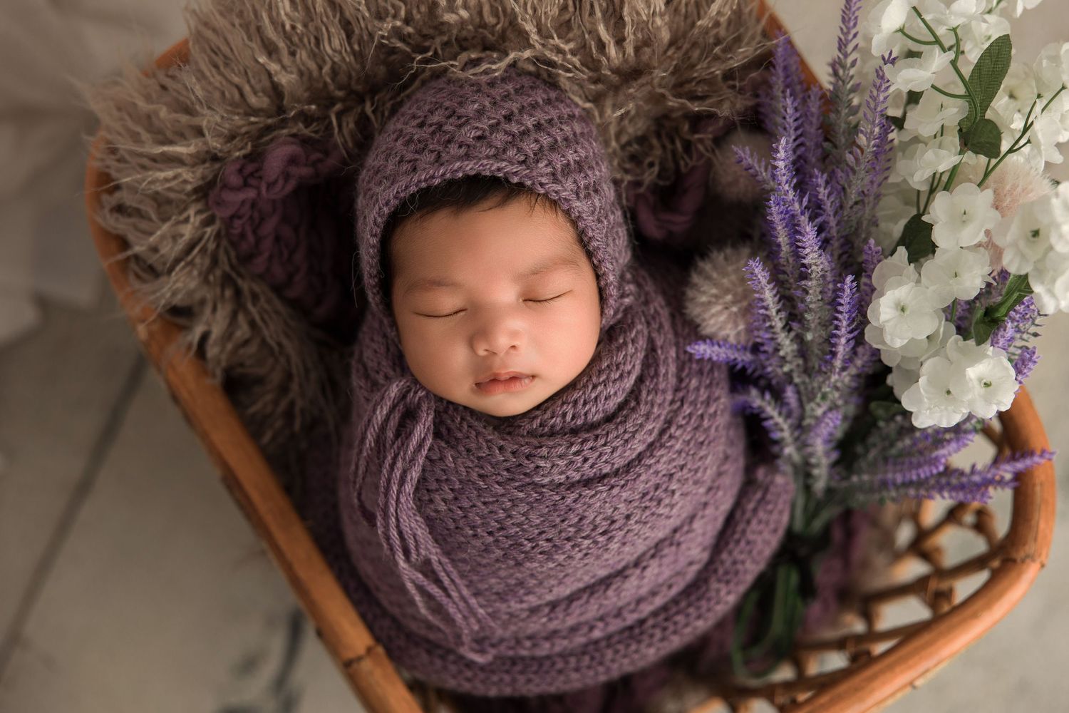 Sleeping newborn wrapped in purple knit blanket rests in wooden bowl with floral decorations and fur lining.