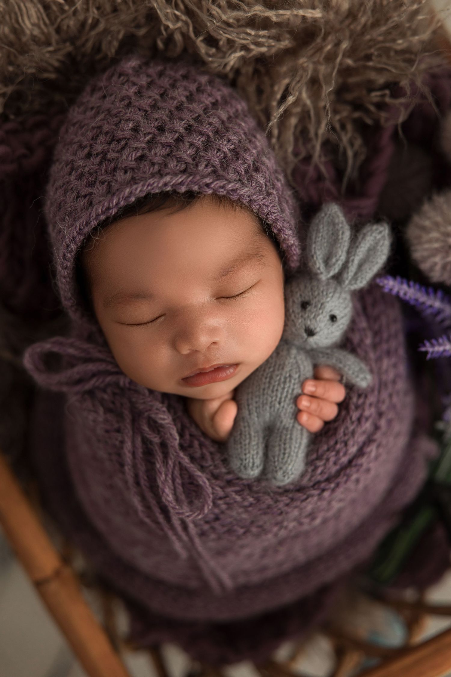 Sleeping baby wrapped in purple knit blanket holding small gray stuffed bunny toy.