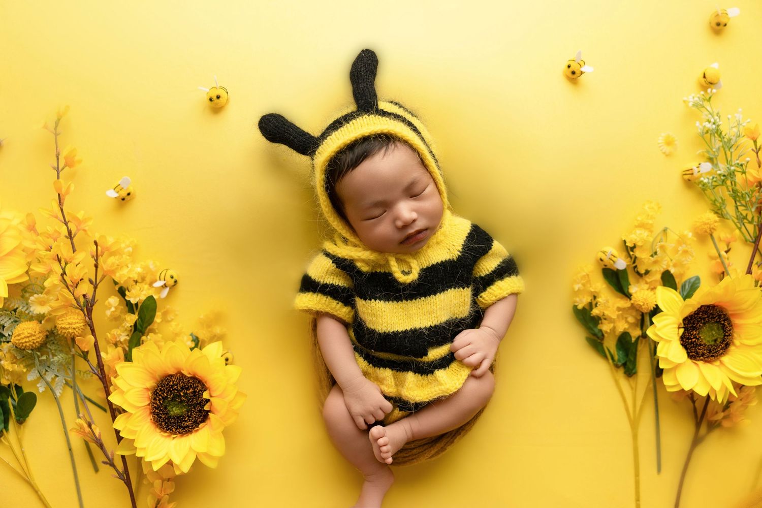 A baby dressed in a yellow and black striped bumblebee costume lies next to sunflowers on a yellow background.