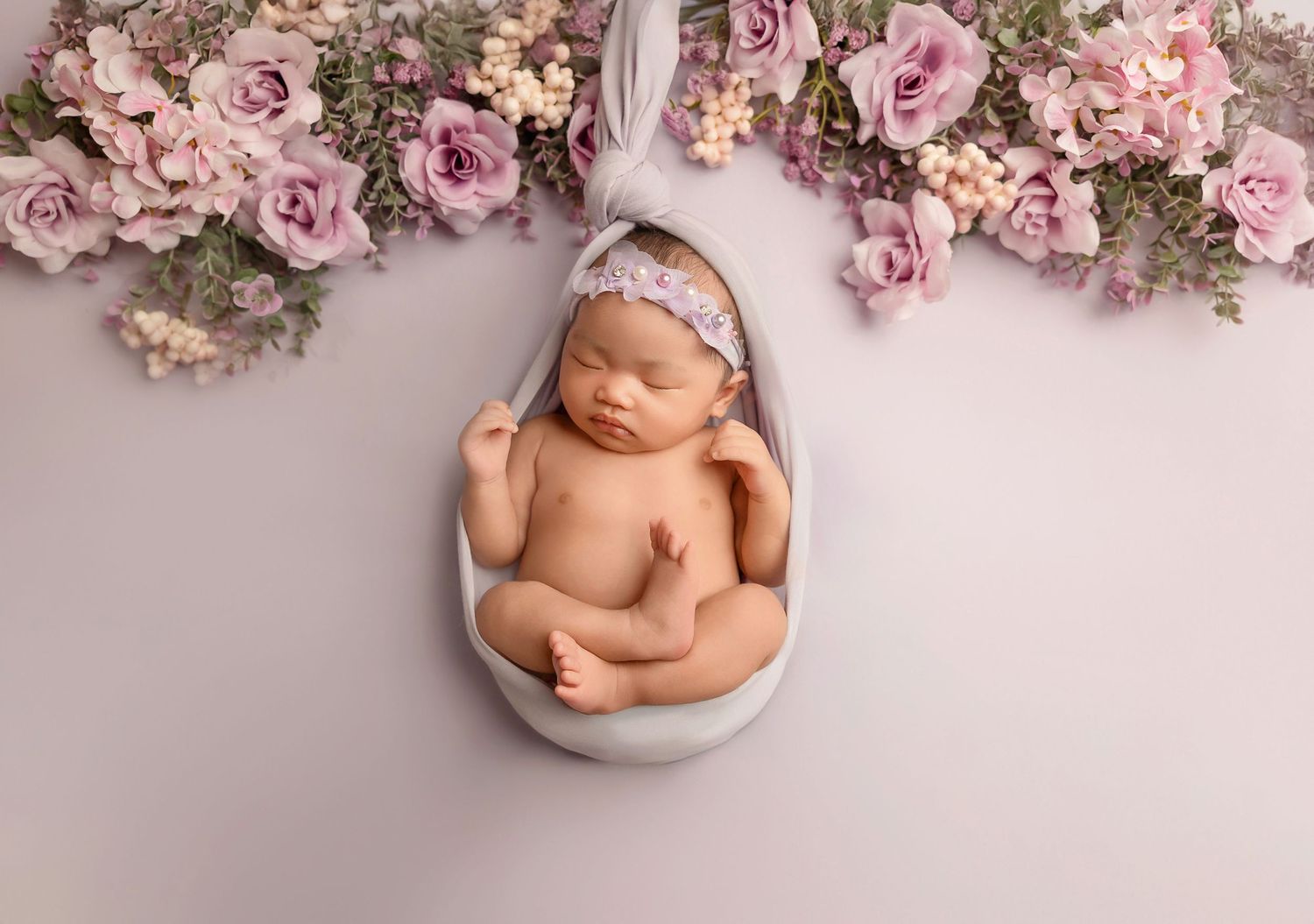 Delicate pink roses and floral arrangements surround a sleeping newborn in a white bowl against a soft pink backdrop.