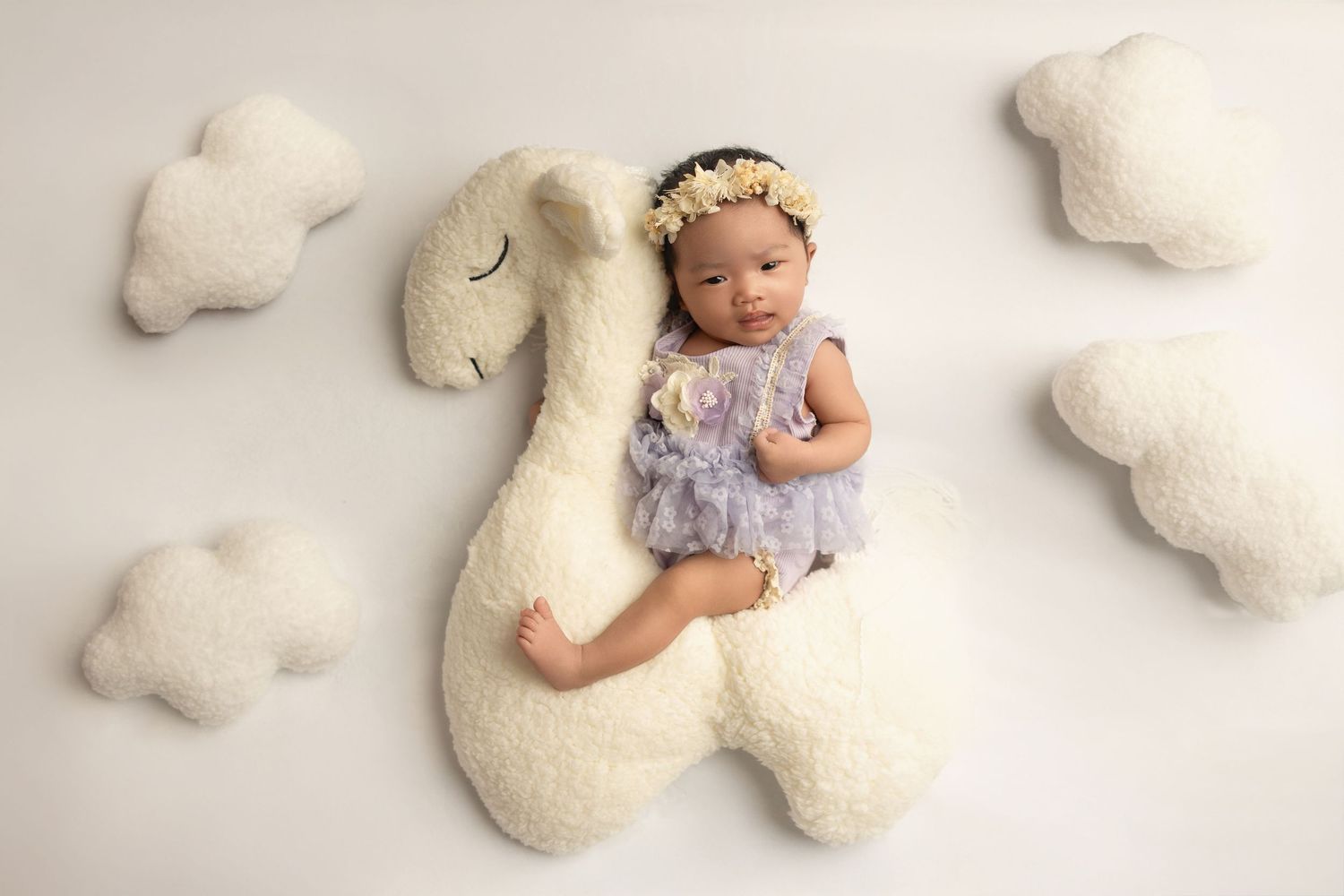Adorable baby wearing purple lace dress lying on white fluffy blanket surrounded by plush teddy bears.