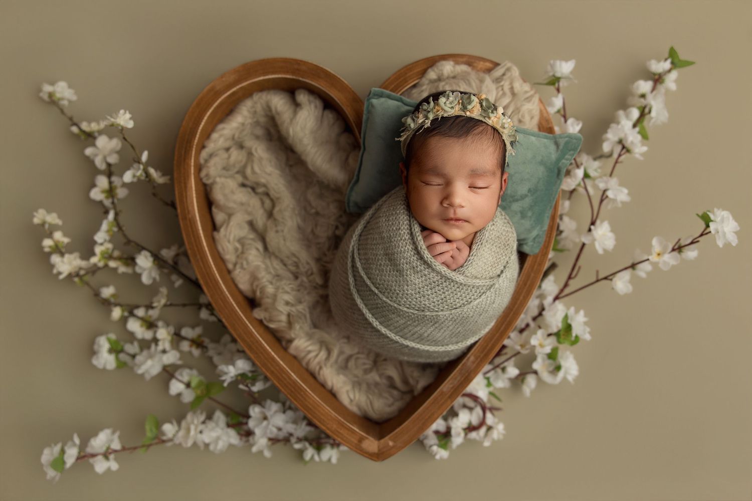 Newborn baby swaddled in green wrap sleeping in heart-shaped wooden bowl surrounded by white cherry blossoms.