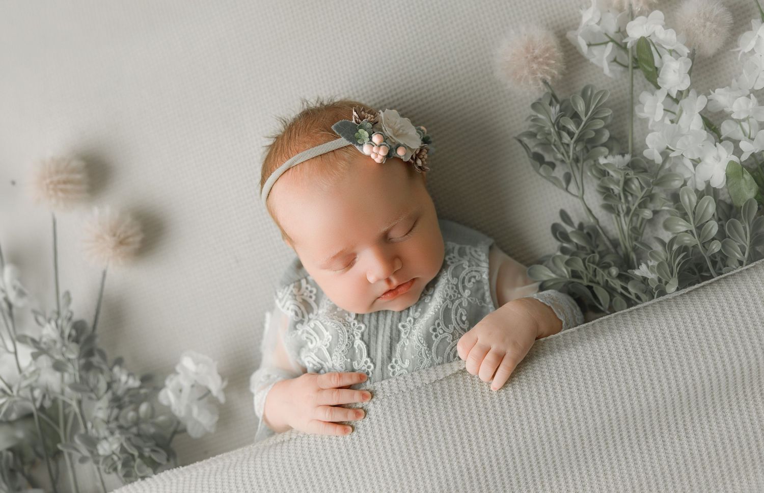 A baby lies on a grey blanket surrounded by soft white floral decorations in a gentle newborn portrait setting.