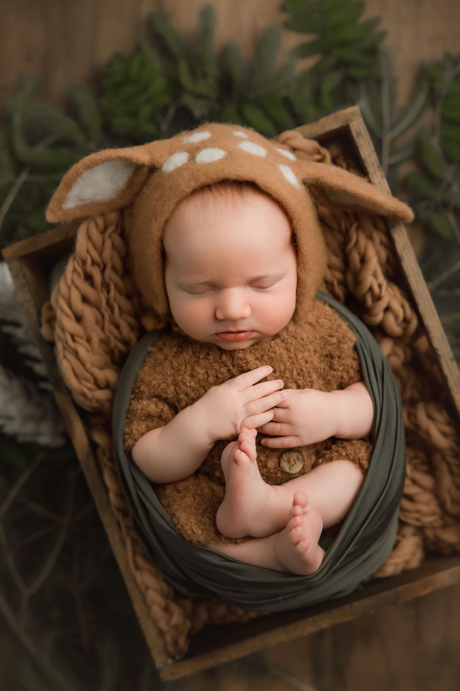 A sleeping baby wearing a brown knit hat lies peacefully in a wooden box draped with dark green fabric.