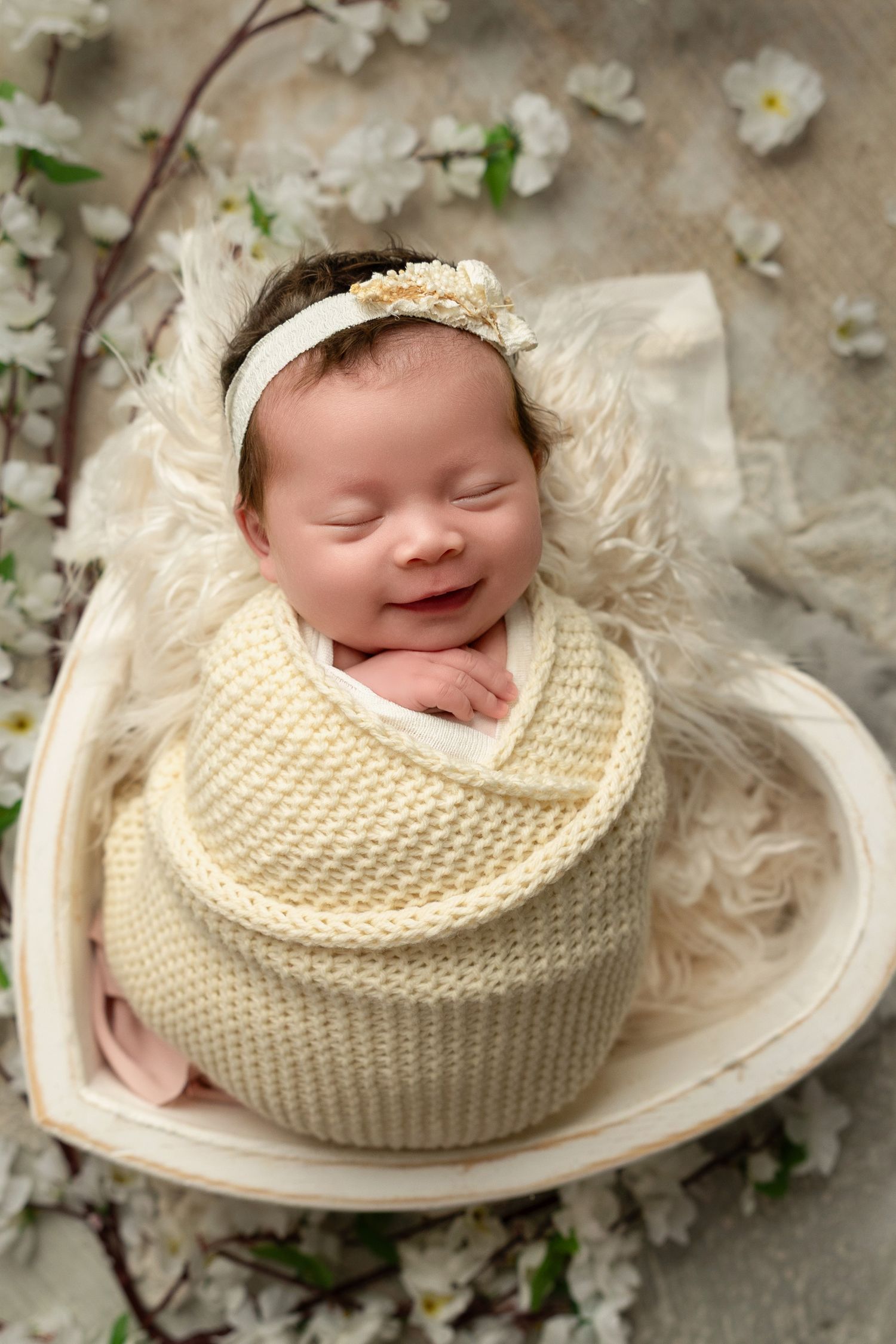Sleeping newborn wrapped in cream knit blanket lies in heart-shaped basket surrounded by delicate white spring blossoms.