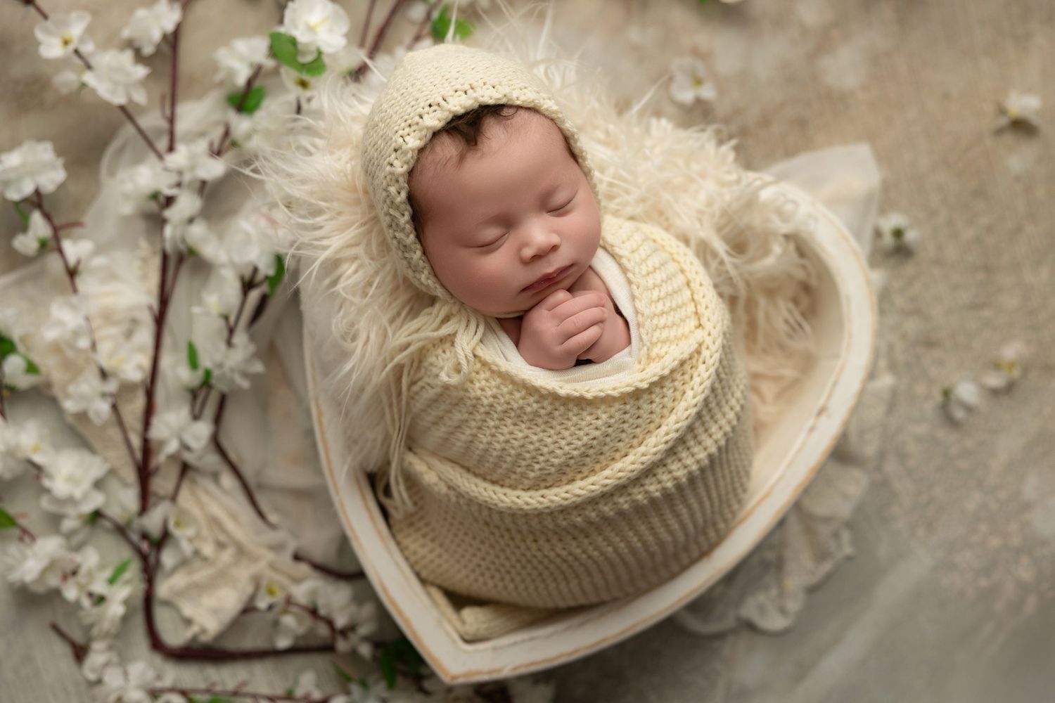 Newborn baby sleeping peacefully in cream knit wrap surrounded by delicate white cherry blossoms.