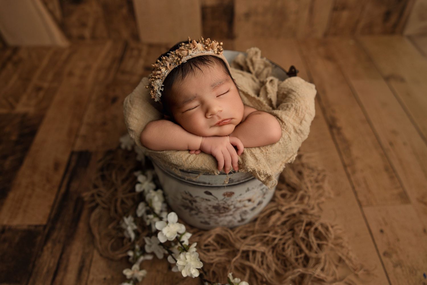 Sleeping newborn baby in a rustic bucket with floral crown and white flowers on a wooden floor backdrop.