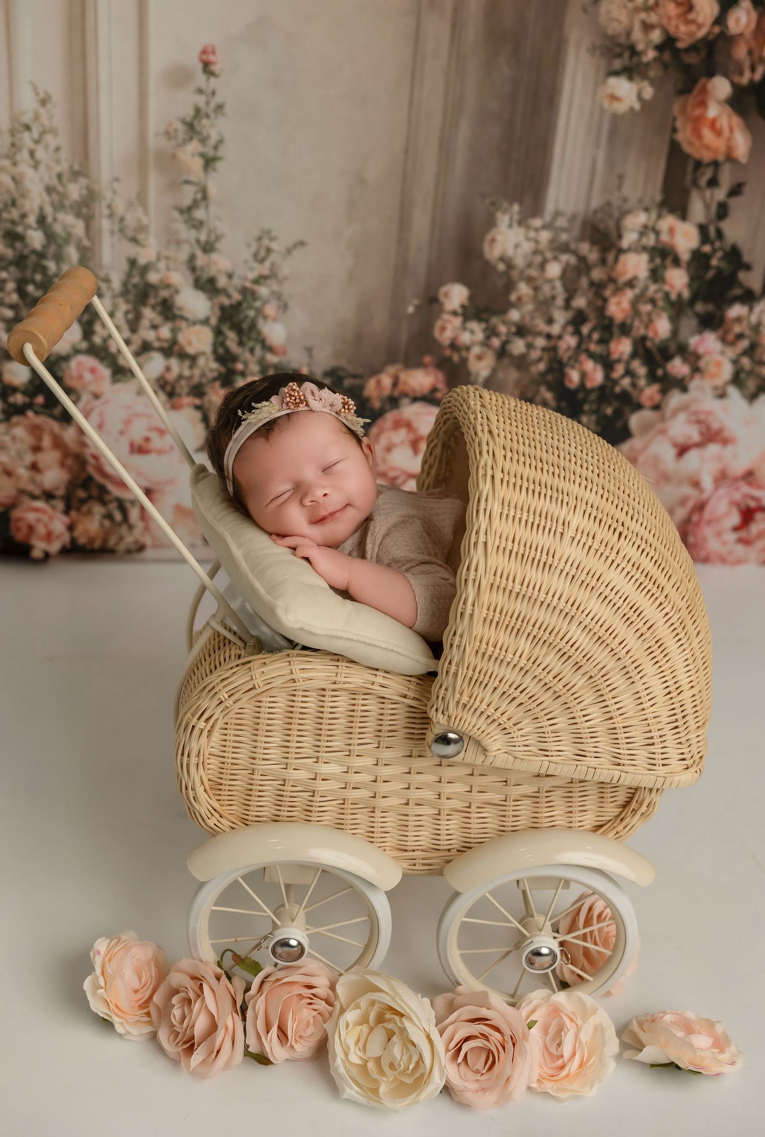 A vintage-style wicker baby carriage decorated with soft pink roses against a neutral backdrop with floral accents.
