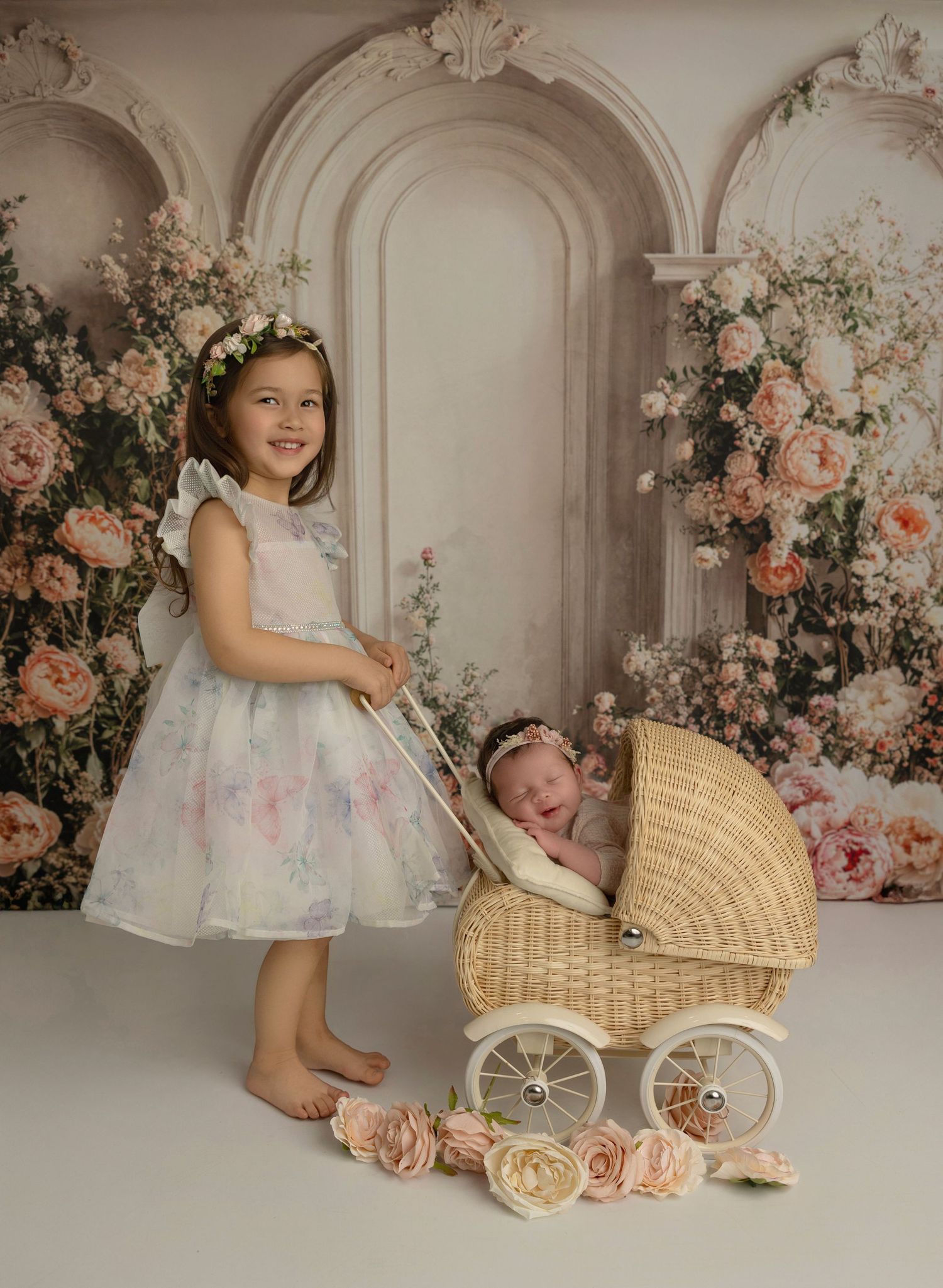 Vintage-style portrait with floral arches, white dress, and wicker baby carriage surrounded by pink roses and decor.