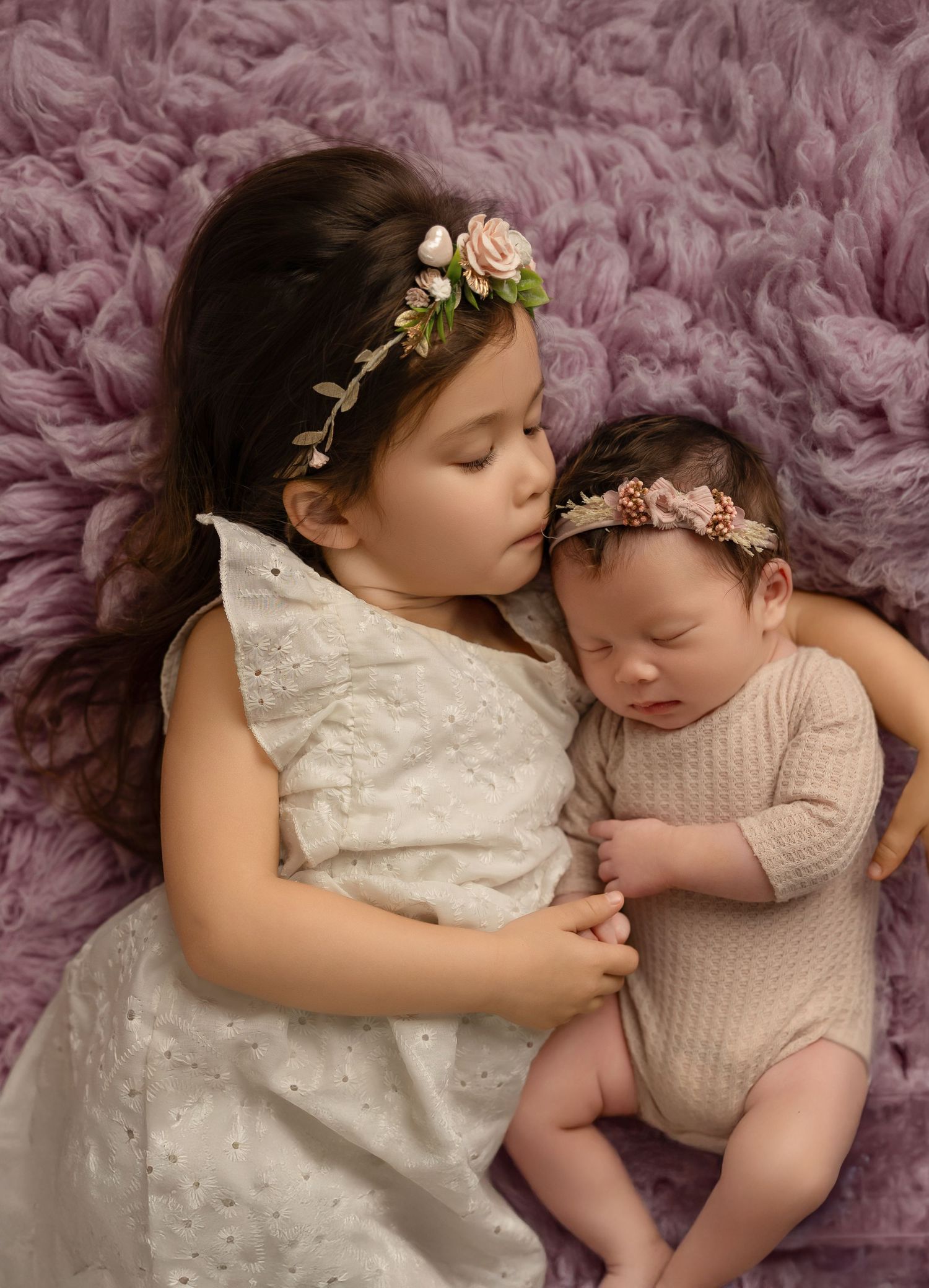 Two siblings in white dresses lie together on a pink furry backdrop during a professional photo session.