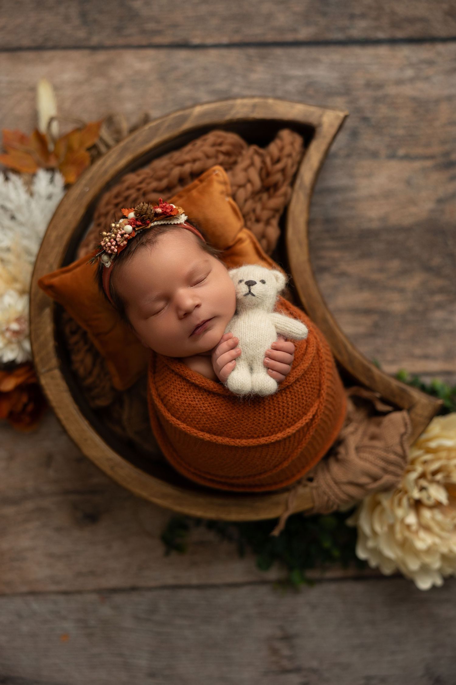 A sleeping newborn in a rustic wooden bowl wearing a brown knit wrap and holding a small white teddy bear.